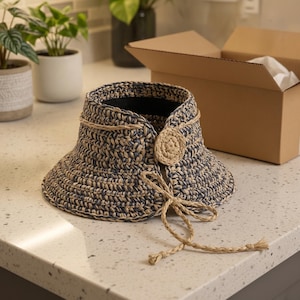 Puede incluir: Una visera de sol tejida a ganchillo con una mezcla de fibras azul marino oscuro y color canela natural. La visera presenta un ala ancha y estructurada, un detalle de botón circular de ganchillo beige y finos lazos trenzados.