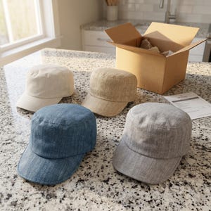Könnte beinhalten: Vier militärische Cadet-Caps in Creme, Beige, Blau und Grau, jeweils mit flacher Oberseite, kurzem gebogenem Schirm und strukturierter Stoffoberfläche. Die Kappen sind gruppiert, mit einem offenen Versandkarton aus Pappe im Hintergrund.