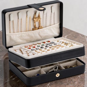 May include: A black rectangular jewelry box with a cream-colored interior. The lid features hooks for necklaces and a pouch. The top tray contains slots for rings and small earrings, while the bottom drawer has divided compartments for storage.