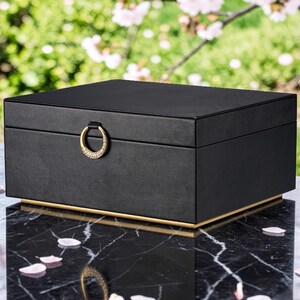 May include: A rectangular black jewelry box with a smooth matte finish, featuring a gold-toned circular ring handle on the front lid and a thin gold-toned base trim. The box has a clean, structured design with sharp edges and a solid black exterior.
