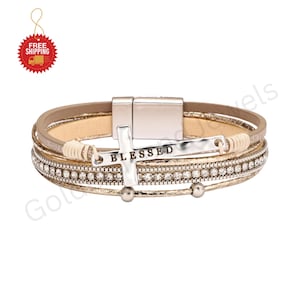 May include: A multi-strand bracelet featuring a silver-toned metal cross charm with the word BLESSED engraved on it. The bracelet includes layers of beige leather, a row of clear rhinestones, and a metallic clasp closure with a magnetic mechanism.