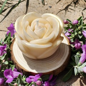 Puede incluir: Una vela con forma de rosa de color crema con bordes de pétalos espolvoreados en oro y una mecha blanca central, apoyada sobre una rodaja de madera circular. La vela presenta pétalos esculpidos en capas que imitan una flor en plena floración.