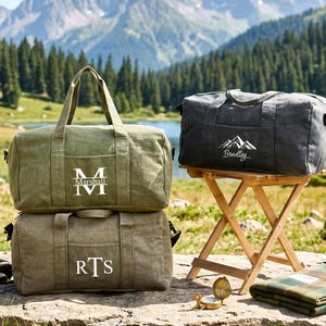 May include: Two olive green canvas duffel bags stacked vertically, one featuring the monogram M and the name Marshall, the other with the initials RTS. A third charcoal gray duffel bag with a mountain graphic and the name Bradley sits on a wooden stool.