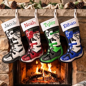 Puede incluir: Cuatro calcetines navideños con forma de botas de motocross, cada uno con un puño blanco con nombres bordados: Jacob en gris, Noah en rojo, Tyler en verde y Ethan en azul. Las botas presentan detalles en colores negro, blanco, rojo, verde y azul.