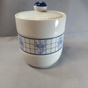 Op de afbeelding: Een witte keramische suikerpot met een deksel voorzien van een donkerblauwe knop. Het lichaam is versierd met een horizontale band van blauwe rasterpatronen met illustraties van bloemstukken in kannen, omrand door dunne blauwe lijnen.
