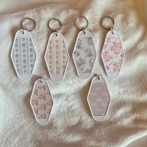 May include: Six diamond-shaped motel-style keyrings with white plastic borders and metal rings. The designs feature various floral and ribbon patterns in shades of blue, pink, and beige, arranged in a grid of four on top and two below.