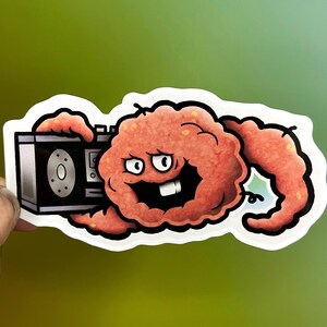 May include: A die-cut sticker featuring a cartoonish, textured orange creature with a wide, toothy grin holding a black boombox. The character has a rounded body and a long, curved tail, outlined in thick black lines against a white border.