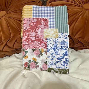 May include: A rectangular fabric-covered journal featuring a patchwork design of various floral, gingham, and striped patterns in shades of red, blue, green, yellow, and white. The cover includes visible stitching and a gold-toned metal charm attached.