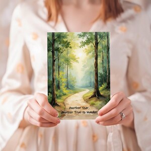 May include: A greeting card featuring a watercolor painting of a forest path leading towards a bright, sunlit clearing. The card has the text "Another Year, Another Trail to Wander". The artwork is predominantly green and yellow.