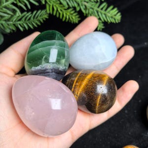 May include: Four polished, egg-shaped stones resting in a palm. The collection includes a translucent green and purple banded stone, a pale blue stone, a light pink stone, and a brown and gold striped tiger's eye stone.