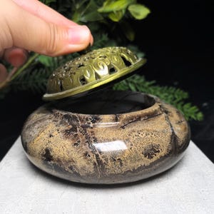 May include: A round, polished stone incense burner with a mottled tan and dark brown pattern. The burner features a removable, ornate, gold-toned metal lid with intricate cut-out scrollwork patterns, resting partially open on the stone base.