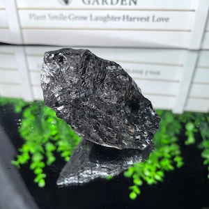 May include: A raw, jagged piece of black tourmaline crystal with a highly reflective, metallic luster. The stone features a rough, uneven surface with deep ridges and sharp edges, displaying a dark, opaque color throughout its crystalline structure.