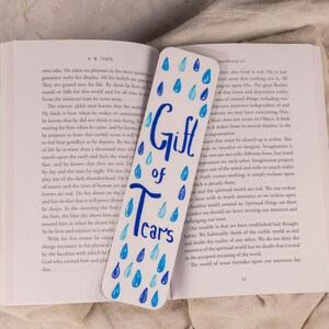 May include: A white rectangular bookmark featuring the blue hand-lettered text Gift of Tears, surrounded by various shades of blue painted raindrops. The bookmark is placed vertically across the open pages of a book with black printed text.