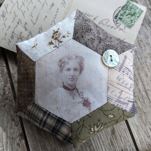 Puede incluir: Un cojín de alfileres hexagonal hecho de tela de patchwork con una fotografía vintage de una mujer en el centro. La tela es una mezcla de patrones florales, cuadros y un diseño de nota musical. Un botón blanco está cosido en el lateral del cojín de alfileres.