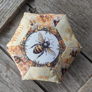 May include: Hexagonal fabric item with a bee design. The central image is a detailed bee illustration, encircled by floral and honeycomb patterns. The fabric uses a light yellow and beige colour scheme, with additional bee motifs on the edges.