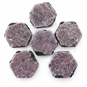 May include: Six hexagonal raw ruby crystals arranged in a circular pattern. Each stone features a flat, textured, dusty-pink surface with visible crystalline structures and dark, rough, black mineral edges surrounding the perimeter of the shapes.