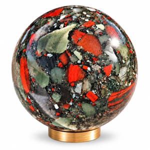 Op de afbeelding: Een gepolijst bolvormig stenen ornament met een complex mozaïek van rode, donkergroene, zwarte en grijze mineraalfragmenten. De bol rust op een ronde voet van geborsteld goudkleurig metaal en heeft een glad, reflecterend oppervlak.