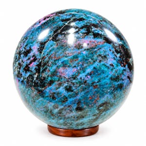Op de afbeelding: Een gepolijste stenen bol met een levendige mix van blauwgroene, blauwe en zwarte mineraalinsluitingen met verspreide vlekken van felroze. De bol rust op een kleine, ronde, donkerbruine houten standaard en heeft een glad, reflecterend oppervlak.