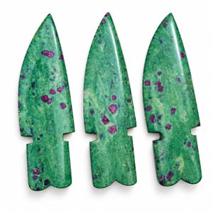 May include: Three polished stone arrowheads carved from green zoisite with embedded magenta ruby crystals. Each piece features a smooth, tapered blade shape with a notched base, showcasing a natural mottled texture and vibrant color contrast.