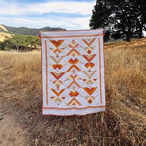 May include: A rectangular quilt featuring a geometric pattern of triangles and diamonds in shades of orange, yellow, and white. The quilt has a solid orange border and is constructed with visible stitching throughout the fabric surface.
