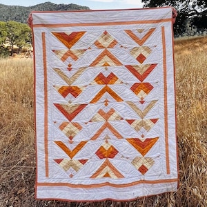 May include: A rectangular quilt featuring a geometric pattern of triangles and chevrons in shades of white, cream, light orange, and burnt orange. The quilt has a solid white border with thin orange stripes and visible linear quilting stitches.