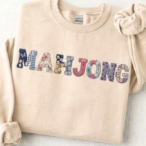 Puede incluir: Una sudadera de cuello redondo en color beige con la palabra MAHJONG aplicada en el pecho. Cada letra está confeccionada con diferentes telas estampadas, incluyendo cuadros vichy azules, motivos florales, mariposas y fichas de mahjong.