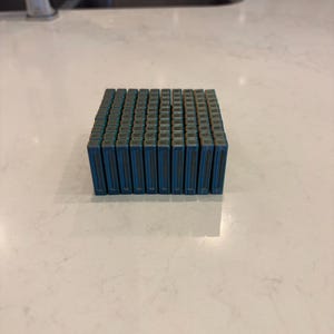 Peut inclure: Une grille carrée de dix par dix petits blocs rectangulaires aux tons bleus, disposés en une formation compacte. Chaque bloc individuel présente une surface texturée à l'aspect métallique avec une rainure verticale distincte au centre.