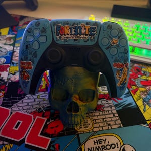 May include: A light blue video game controller featuring various Fortnite-themed stickers and the text "FORTNITE" and "WHERE WE DROPPIN" resting on a yellow and blue skull-shaped stand. The controller has black joysticks and buttons.