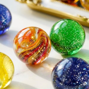 Puede incluir: Una colección de canicas de vidrio esféricas con varios diseños. Una canica muestra patrones arremolinados de color naranja, rojo y amarillo con purpurina incrustada, mientras que otras son de color verde o azul sólido con partículas brillantes.