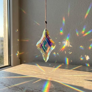 May include: A faceted, diamond-shaped glass crystal suncatcher hangs from a thin brown cord. The clear glass prism refracts sunlight, casting vibrant, multi-coloured rainbow light patterns across the surrounding area and the surface below.