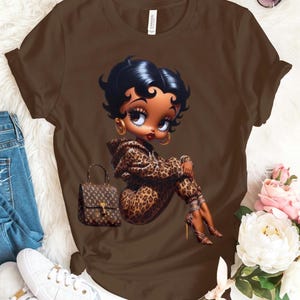 May include: A brown short-sleeved t-shirt featuring a graphic of a cartoon character with black hair wearing a leopard print hooded outfit and high heels, accompanied by a brown handbag with a repeating logo pattern printed on the front.