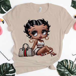 May include: A beige short-sleeved t-shirt featuring a graphic of a cartoon character with black hair, large eyes, and gold hoop earrings. The character wears a white outfit with red and green stripes and the text GUCCI, sitting beside a handbag.