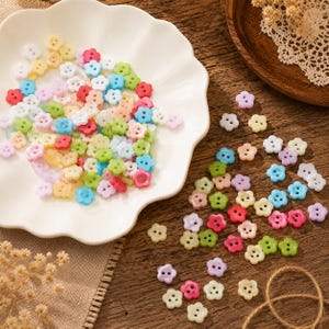 Puede incluir: Una colección de pequeños botones de plástico multicolor con forma de flores de cinco pétalos. Los botones tienen dos agujeros en el centro y vienen en varios colores pastel y brillantes, incluyendo rosa, azul, verde, amarillo, blanco y morado.