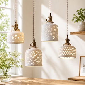 May include: Four white ceramic pendant lights with intricate cut-out patterns, including circles, flowers, and geometric grids. Each shade is suspended by a twisted brown cord from a brass-toned metal fixture, creating a warm, illuminated display.