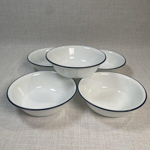 May include: A set of four white enamel bowls with a dark blue rim. The bowls are arranged in a cluster, featuring a smooth, glossy finish and a simple, rounded design. Each bowl has a consistent shape and color, creating a uniform appearance.
