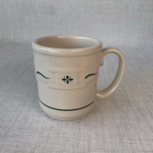 May include: A cream-colored ceramic mug featuring a dark green embossed floral motif at the center, flanked by two curved horizontal lines. The mug has a sturdy handle on the right side and a single dark green horizontal line near the base.