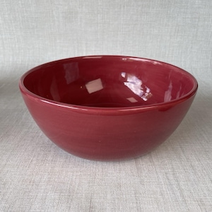 May include: A deep, round ceramic bowl with a smooth, glossy finish in a solid dark red color. The bowl features a wide, open top and a curved, uniform shape, reflecting light on its interior surface, showcasing a simple and functional design.