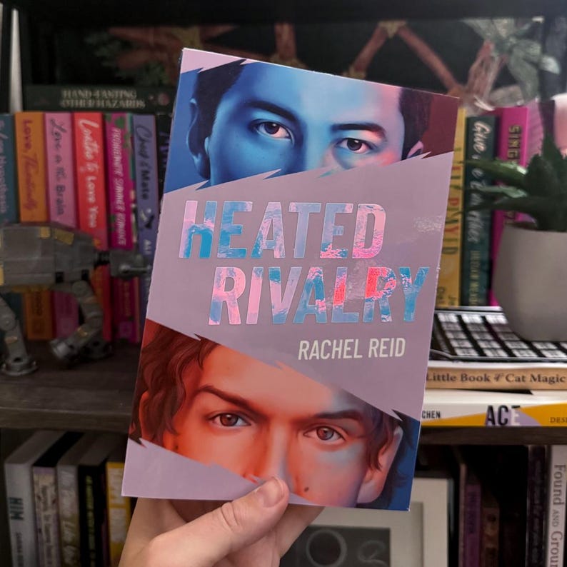 May include: A paperback book titled Heated Rivalry by Rachel Reid. The cover features a split design with a person's eyes in blue tones at the top and another person's eyes in warm tones at the bottom, separated by a jagged white diagonal line.