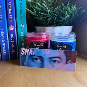 May include: Two glass jars with pink and blue wax, each featuring a black label with the text Briarwick. In front, a rectangular card displays the text SHANE 24 above a detailed illustration of a pair of eyes with blue and purple tones.