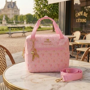 May include: A light pink lunch bag with a floral pattern, featuring the Stanley and LoveShackFancy logos in gold text. The bag has two top handles, a front zippered pocket, and a detachable pink shoulder strap with gold-tone metal hardware.