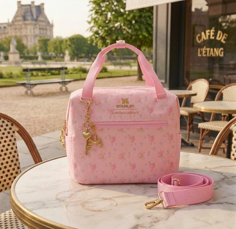 May include: A light pink lunch bag with a floral pattern, featuring the Stanley and LoveShackFancy logos in gold text. The bag has two top handles, a front zippered pocket, and a detachable pink shoulder strap with gold-tone metal hardware.
