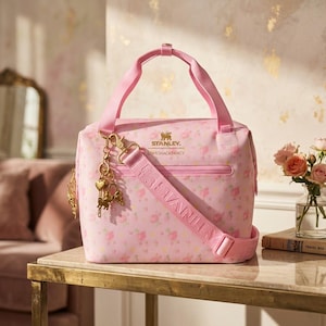 May include: A light pink lunch bag featuring a floral pattern, two top handles, and an adjustable shoulder strap. The front displays the gold Stanley and LoveShackFancy logos, with a gold-toned charm attached to the strap hardware.