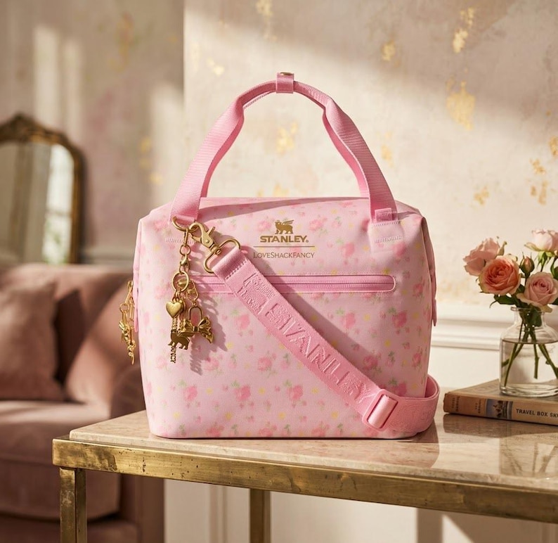May include: A light pink lunch bag featuring a floral pattern, two top handles, and an adjustable shoulder strap. The front displays the gold Stanley and LoveShackFancy logos, with a gold-toned charm attached to the strap hardware.