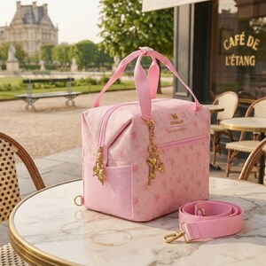 May include: A light pink lunch bag featuring a floral pattern, two top handles, and a detachable shoulder strap. The bag displays the Stanley logo and a gold-toned charm set with a heart and bow. It includes a front zippered pocket and side pockets.
