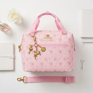 May include: A light pink lunch bag featuring a floral pattern, gold-tone hardware, and a gold charm set with a heart, a winged horse, and a circular emblem. The bag displays the Stanley and LoveShackFancy logos in gold text and includes a detachable strap.