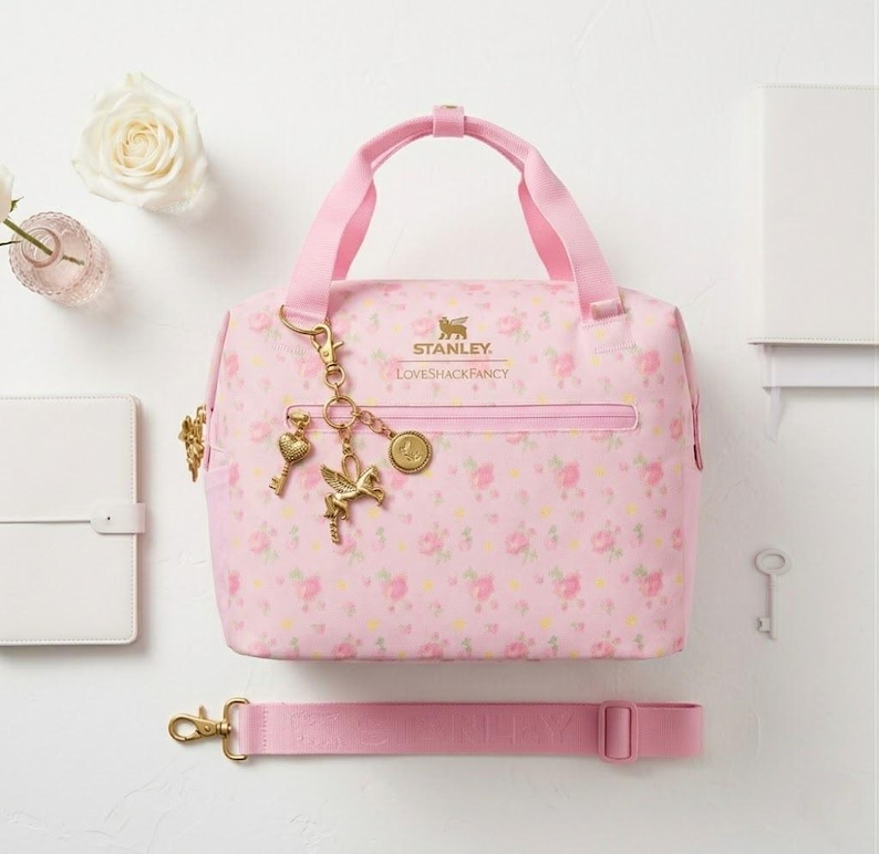 May include: A light pink lunch bag featuring a floral pattern, gold-tone hardware, and a gold charm set with a heart, a winged horse, and a circular emblem. The bag displays the Stanley and LoveShackFancy logos in gold text and includes a detachable strap.