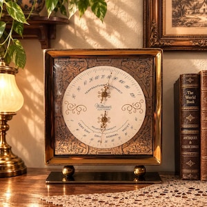 May include: A square Wittnauer Humi-Therm instrument with a gold-toned frame and two circular dials measuring temperature in Fahrenheit and relative humidity. The device features ornate decorative patterns and stands on two small gold spheres.