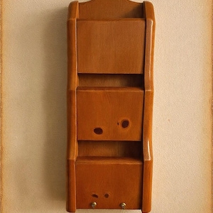 May include: A vertical wooden wall organiser featuring two stacked storage compartments with a smooth, medium-brown finish. The structure includes a curved top edge and two small metal hooks attached to the bottom front for hanging items.