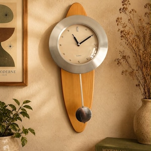 May include: A wall clock featuring a circular brushed silver metal frame surrounding a cream-coloured face with black hands and small silver dot markers. The clock is mounted on a vertical, light-toned wooden surfboard-shaped backing with a silver pendulum.
