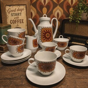 May include: A white ceramic tea set featuring a repeating orange, yellow, and brown mandala-style floral pattern. The set includes a tall teapot with a lid, a creamer, a lidded sugar bowl, and several teacups with matching saucers.